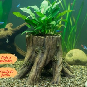 Puede incluir: Un adorno de acuario con forma de tocón de árbol marrón texturizado con un centro hueco que contiene una planta acuática verde. El adorno presenta detalles realistas de corteza y raíces. Dos etiquetas de texto naranja a la izquierda dicen Prêt à peindre y Ready to paint.