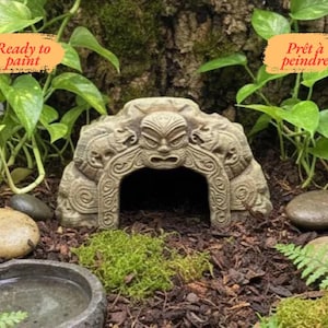 Puede incluir: Un escondite decorativo, listo para pintar, con forma de piedra, con un diseño de rostro tallado, rodeado de vegetación y rocas. La imagen incluye el texto "Ready to paint" y "Prêt à peindre".