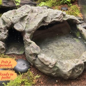 Può includere: Un rifugio per rettili e abbeveratoio dalla texture rocciosa grigia, dotato di un ingresso a grotta e di una vaschetta poco profonda con un motivo topografico. L'oggetto include un'etichetta arancione con il testo "Prêt à peindre" e "Ready to paint".