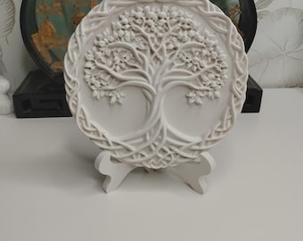 May include: A white, circular relief sculpture of a tree with intricate details, set on a white stand. The tree design is surrounded by a Celtic knot border. The sculpture is displayed against a blurred background.