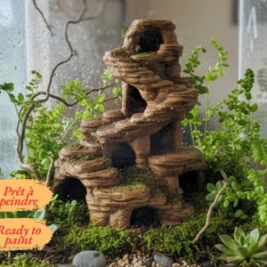 May include: A brown, multi-level rock-like structure with several cave openings and textured surfaces, surrounded by green ferns and succulents inside a glass terrarium. The text "Prêt à peindre" and "Ready to paint" is visible on orange labels.