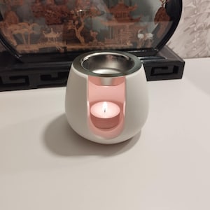 Peut inclure: Brûle-parfum en céramique blanche avec un dessus argenté et une petite bougie à l'intérieur. Le brûleur a un intérieur rose et une forme ronde.