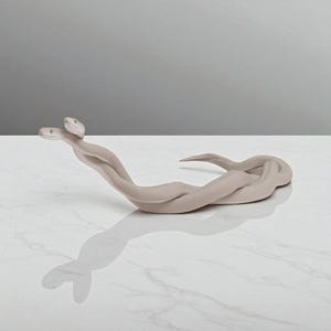 Puede incluir: Una escultura beige de dos serpientes entrelazadas. Las serpientes están anudadas, con sus cabezas levantadas y mirando en la misma dirección. La escultura está sobre una superficie de mármol blanco.