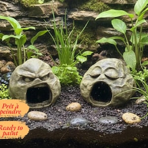 May include: Two gray, rock-shaped resin hides with carved facial expressions, one with an angry face and one with a sleeping face, each featuring a hollow opening. The text "Ready to paint" is printed on an orange background in the corner.