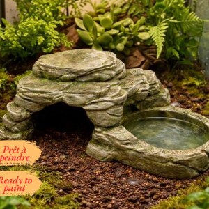 Könnte beinhalten: Ein graues, felsförmiges Reptilienversteck mit integrierter Wasserschale, strukturierter Oberfläche und einem hohlen Bogeneingang. Das Objekt enthält zwei orangefarbene Etiketten mit dem in Schwarz gedruckten Text "Prêt à peindre" und "Ready to paint".