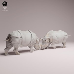 Peut inclure: Deux figurines de rhinocéros blancs en position de combat. Le rhinocéros de gauche a un design de corps segmenté. Les rhinocéros se font face, leurs cornes se touchant presque. L'arrière-plan est de couleur neutre unie.