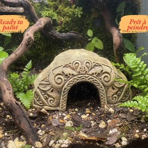 Könnte beinhalten: Beige, dekorative höhlenförmige Terrariendekoration mit aufwendigen Schnitzereien. Das Ornament hat einen bogenförmigen Eingang und ist von Grün und kleinen Steinen umgeben. Die Worte "Prêt à peindre" sind auf einem gelben Etikett zu sehen.