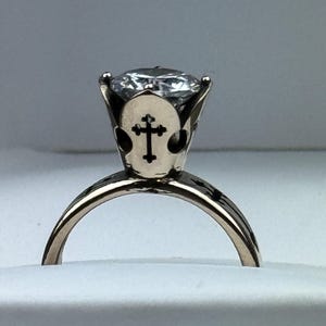 May include: A gold ring with a large, clear gemstone. The setting has a cross design on the sides. The ring is set against a white background, highlighting its details and craftsmanship. A piece of jewellery.