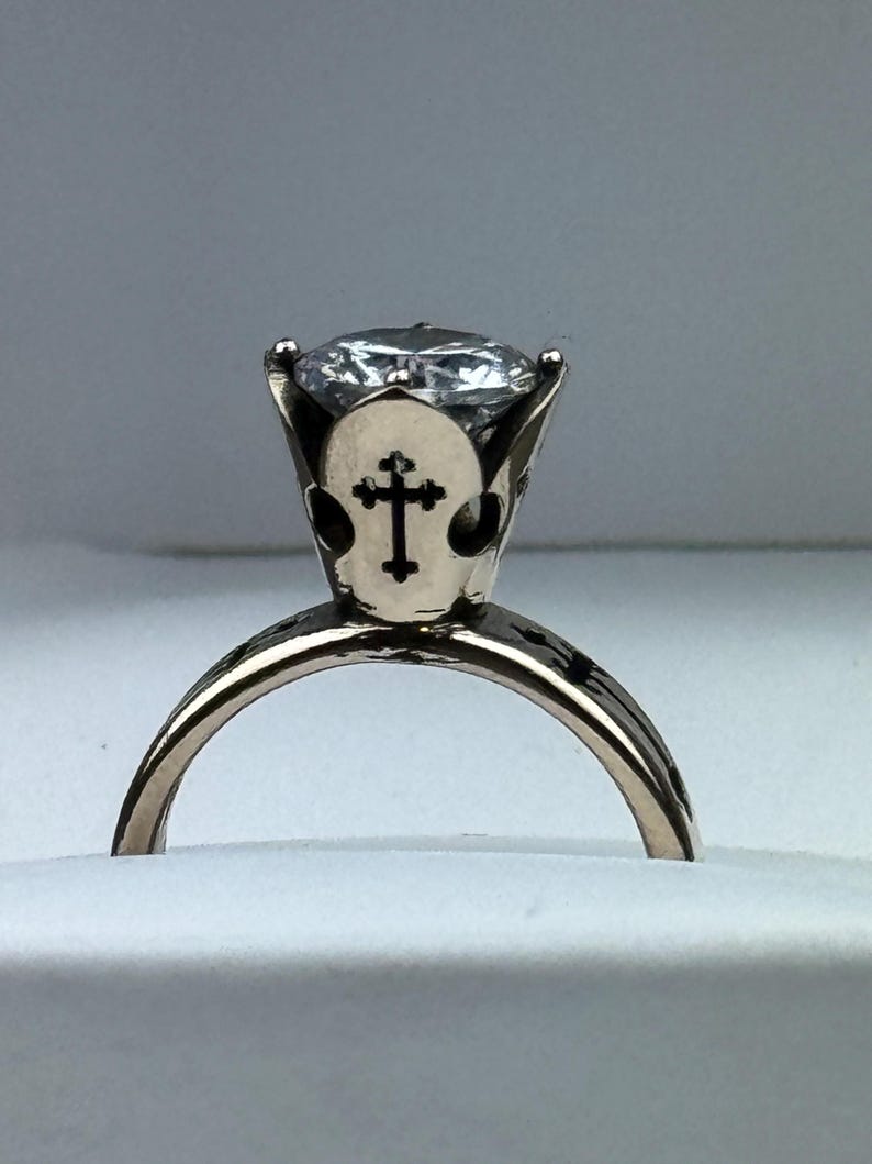 May include: A gold ring with a large, clear gemstone. The setting has a cross design on the sides. The ring is set against a white background, highlighting its details and craftsmanship. A piece of jewellery.