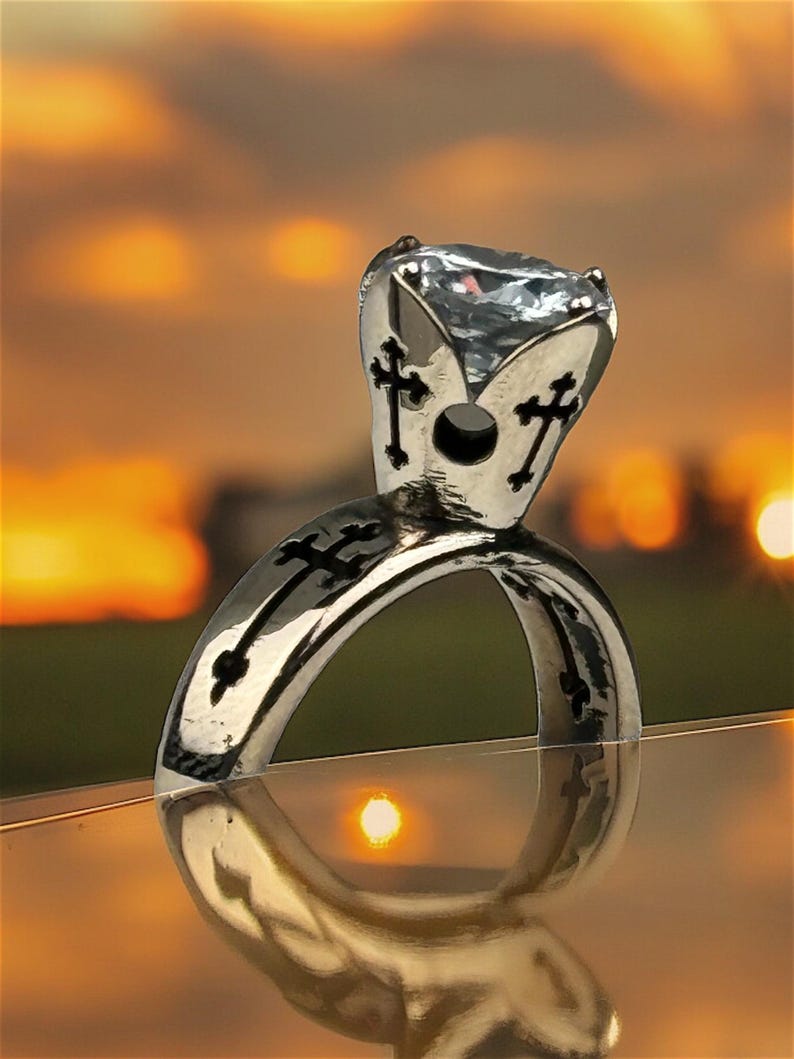 May include: A silver ring with a large, clear gemstone. The ring has a cross design on the band and around the gemstone setting. The ring is highly reflective, with a bright, polished finish.