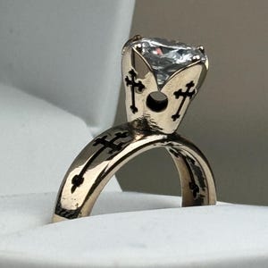 May include: A gold-coloured ring with a large, clear gemstone. The ring features a cross design on the band and around the gemstone setting. The ring is displayed in a white ring box.