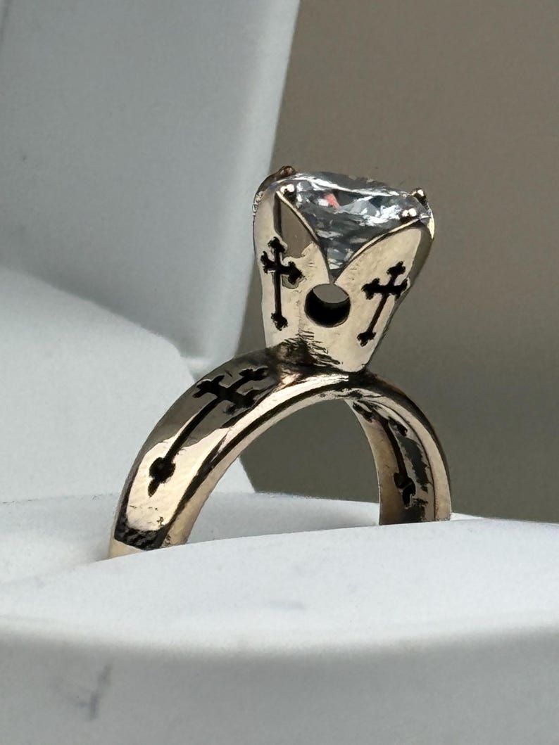 May include: A gold-coloured ring with a large, clear gemstone. The ring features a cross design on the band and around the gemstone setting. The ring is displayed in a white ring box.
