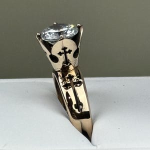 May include: A gold ring with a large, clear gemstone. The ring has a cross design on the band and around the gemstone setting. The ring is set against a white background.
