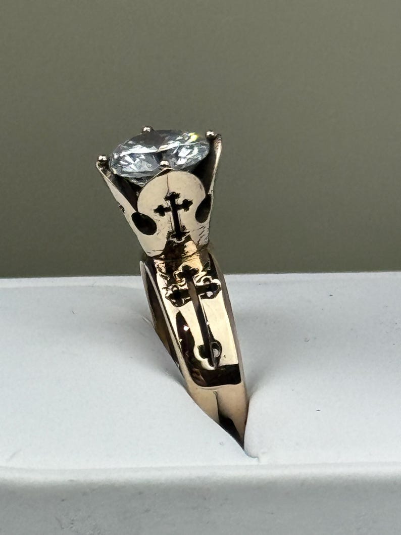 May include: A gold ring with a large, clear gemstone. The ring has a cross design on the band and around the gemstone setting. The ring is set against a white background.