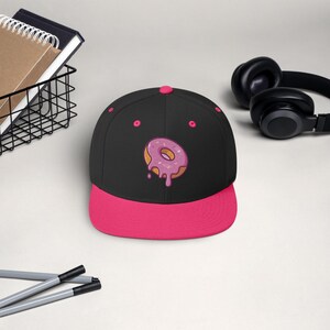 May include: A black and pink baseball cap with a pink and white donut graphic. The donut has pink frosting and sprinkles.