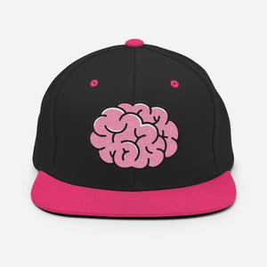Peut inclure: Casquette noire avec un motif de cerveau rose brodé sur le devant. La casquette a une visière rose et un bouton rose sur le dessus. Le motif du cerveau est rose clair avec un contour blanc.