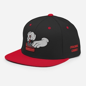 Könnte beinhalten: Schwarze und rote Baseballkappe mit einem gestickten Bild einer Hand, die einen Joystick hält. Der Text "ORIGINAL GAMER" ist an der Seite der Kappe gestickt.