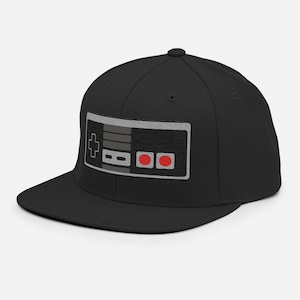 Könnte beinhalten: Eine schwarze und graue Baseballkappe mit einem schwarzen und grauen Videospiel-Controller-Design auf der Vorderseite. Der Controller hat rote Knöpfe.