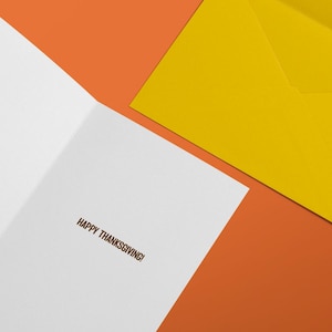 Puede incluir: Una tarjeta de felicitaci&oacute;n blanca con el texto HAPPY THANKSGIVING! impreso en tinta marr&oacute;n en el interior, acompa&ntilde;ada de un sobre amarillo s&oacute;lido, todo colocado sobre un fondo naranja vibrante para crear una presentaci&oacute;n festiva.