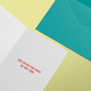 May include: A white greeting card with the red printed text HOPE YOU HAD YOUR SHOTS! GET WELL SOON. The card is positioned next to a solid teal envelope. The items are arranged on a light yellow surface to display the interior message clearly.