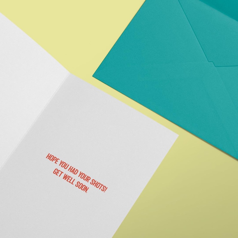 May include: A white greeting card with the red printed text HOPE YOU HAD YOUR SHOTS! GET WELL SOON. The card is positioned next to a solid teal envelope. The items are arranged on a light yellow surface to display the interior message clearly.