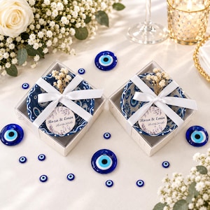 Op de afbeelding: Twee vierkante geschenkdozen met blauw-wit gedessineerde keramische schaaltjes, elk gestrikt met een wit lint, een takje gedroogd gipskruid en een rond label met de tekst Roza & Emin 26.09.2026, omringd door blauwe kralen met een boze oog motief.