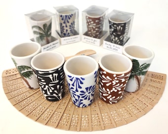 May include: A collection of ceramic shot glasses with various designs, including palm trees and floral patterns in black, blue, and brown. Some glasses are packaged in clear boxes, and are displayed on a decorative fan.