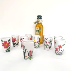 Peut inclure: Un ensemble de neuf verres à liqueur en céramique blanche ornés de divers motifs floraux peints à la main en rouge, rose et vert, disposés autour d'une bouteille en verre remplie d'huile d'olive dorée avec une étiquette portant le texte L'Olio.