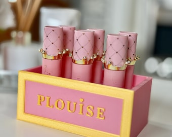 Könnte beinhalten: Eine rosa Lippenstift-Ausstellungsbox mit goldenem Rand und dem Namen "PLOUISE" in goldenen Buchstaben. Die Box enthält mehrere rosa Lippenstifte mit goldenen kronenförmigen Akzenten und Herzmustern.