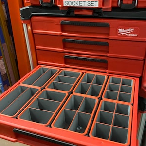 Peut inclure: Boîte à outils roulante Milwaukee Packout rouge avec plusieurs tiroirs. Le tiroir supérieur est ouvert et comporte un insert organisateur gris avec plusieurs compartiments.