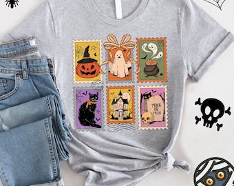 May include: A light gray t-shirt featuring a colorful Halloween-themed design. The design includes postage stamp-style illustrations of a jack-o'-lantern, ghost, cauldron, black cat, haunted house, and a tombstone with the text "Trick or Treat."