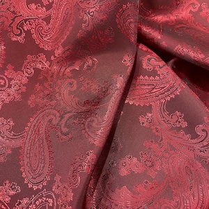 Può includere: Tessuto rosso con motivo paisley e un leggero luccichio. Il tessuto è piegato e drappeggiato, mostrando il design intricato.