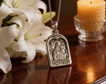 May include: A silver-toned metal pendant necklace featuring a rectangular icon with an arched top. The pendant depicts a religious figure holding a child, with Cyrillic text at the bottom. The piece hangs from a delicate silver-toned chain.