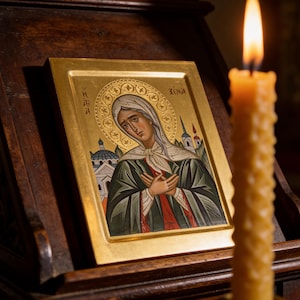 Op de afbeelding: Een religieus icoon met een gouden lijst, dat een figuur met een halo afbeeldt, gekleed in een witte hoofdbedekking en donkergroene gewaden. Het icoon staat op een houten standaard, met een brandende kaars op de voorgrond.