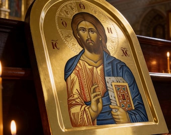 May include: A gold-framed religious icon depicting Jesus Christ, holding a book. The icon features a detailed painting with gold, blue, and red hues. The image is set against a blurred background with lit candles, suggesting a church setting.
