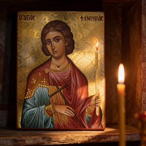 May include: An icon of a saint with a halo, holding a cross and a candle. The icon features a gold background with text in Greek. The saint is depicted in red and blue robes. A lit candle is in the foreground.
