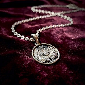 May include: A silver pendant necklace with a detailed religious scene. The round pendant features a raised relief design with figures and architectural elements. The necklace chain is a delicate, linked design, set against a deep burgundy velvet background.