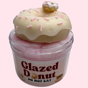 May include: A clear plastic container filled with pink material, topped with a glazed donut and a mini donut. The donut is beige with white icing and pink sprinkles. The container has the words "Glazed Donut DO NOT EAT" printed on it.