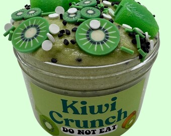 May include: A jar of green slime decorated with kiwi slices, white and black sprinkles, and green chunks. The label reads "Kiwi Crunch" and "DO NOT EAT". The slime is in a clear plastic container.