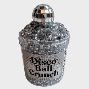 Peut inclure: Un pot en plastique transparent rempli de paillettes argentées et surmonté d'une boule disco argentée. Le pot porte une étiquette noire sur laquelle est inscrit "Disco Ball Crunch" et "Ne pas manger".