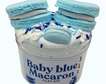 Pode incluir: Um recipiente de plástico transparente cheio de slime branco, coberto com macarons azuis e granulado. O rótulo diz "Baby blue Macaron CLAY KIT". Os macarons são azuis claros com recheio branco.