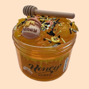 May include: A clear plastic jar filled with yellow slime, decorated with honey-themed embellishments. A wooden honey dipper rests on top. The jar has a label that reads "Honey Cubes".