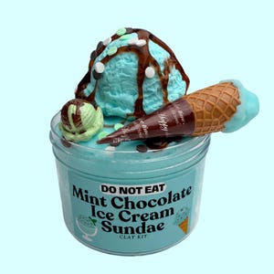 May include: A clear plastic jar filled with light blue slime, topped with a fake ice cream sundae. The sundae includes a scoop of mint green ice cream with chocolate sauce and sprinkles, a scoop of light blue ice cream with chocolate sauce and white sprinkles, and a waffle cone with light blue ice cream. The jar has a label that reads "DO NOT EAT Mint Chocolate Ice Cream Sundae CLAY KIT".