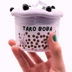 Lavender glossy slime in a clear jar labeled “Taro Boba,” topped with realistic ice cube charms, black boba ball charms, and a mini boba cup. Soft, thick texture and smooth finish, perfect for ASMR and bubble pops. Suitable for slime enthusiasts, collectors, and sensory play lovers.