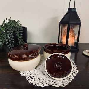 Puede incluir: Cuencos de cerámica marrón y crema de dos piezas con tapas. Una tapa está quitada, revelando el interior y los números "340" y "6816" en la tapa. Una linterna negra, una vela y vegetación están en el fondo.