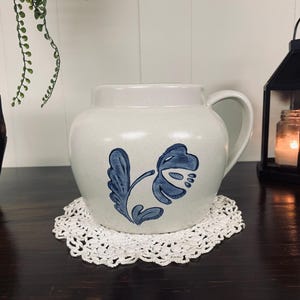 Puede incluir: Una jarra de cerámica blanca con un diseño floral azul. La jarra tiene un asa y una boca ancha.