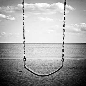 May include: A black and white photo of a swing set against a cloudy sky and a calm ocean. The swing is empty and the chains are hanging down.