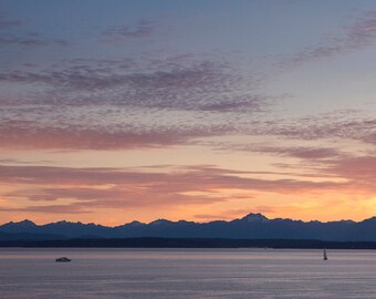 以下が含まれることがあります： 夕焼け空にシルエットになった山脈の雄大な景色。空はピンク、オレンジ、ブルーの色合いで、ふわふわとした雲が広がっています。手前に小さなボートが水面に浮かんでおり、遠くには帆船が見えます。