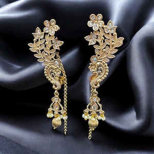 May include: A pair of gold-colored earrings with a floral and leaf design. The earrings feature clear, sparkling accents and dangling chains with small, round beads. The background is a dark, silky fabric.