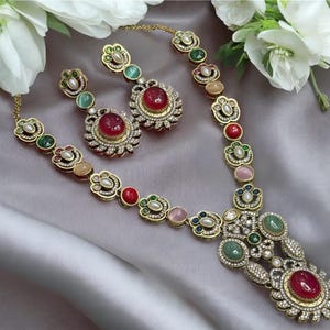 May include: A gold-tone necklace and matching earring set featuring ornate metalwork, clear crystals, and cabochon stones in shades of red, green, and soft pink. The necklace has a central pendant and a chain of alternating floral-shaped links.